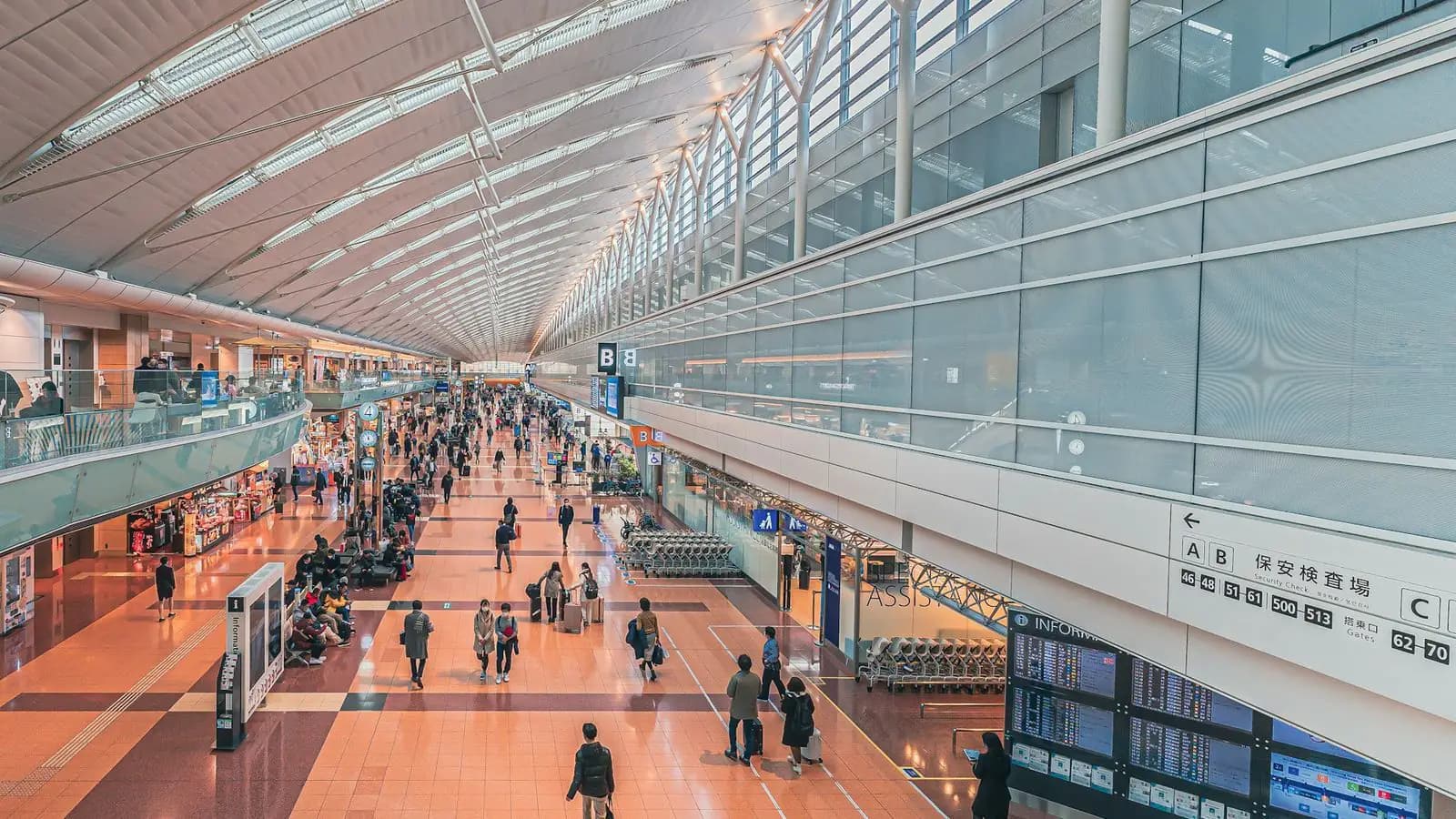 A bright, warm-toned airport terminal hall seen from above, with travellers walking along a long concourse under a curved roof.