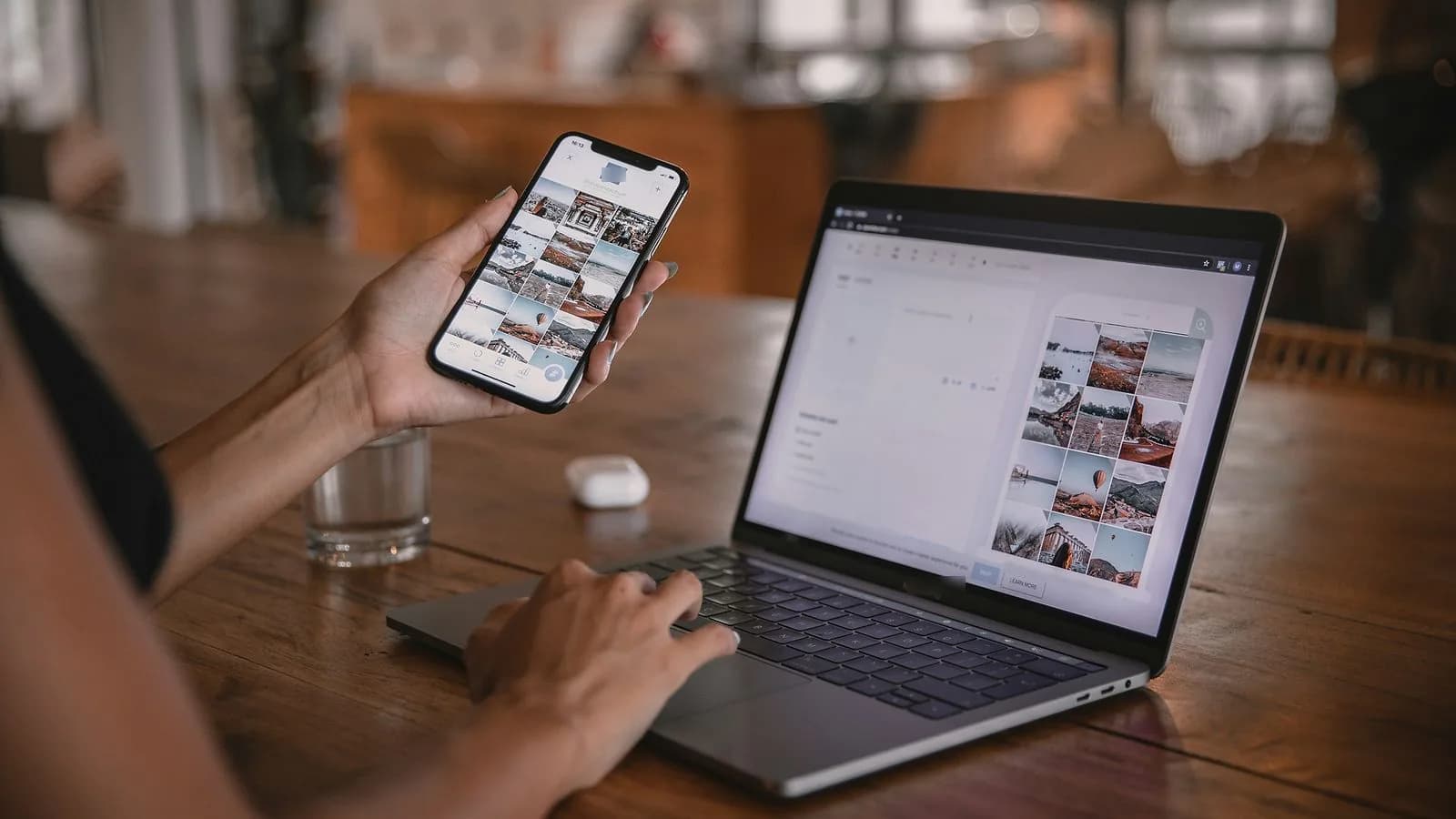 A woman's hands holding a phone and typing on a laptop at a wooden table, both screens showing the same photo gallery page.