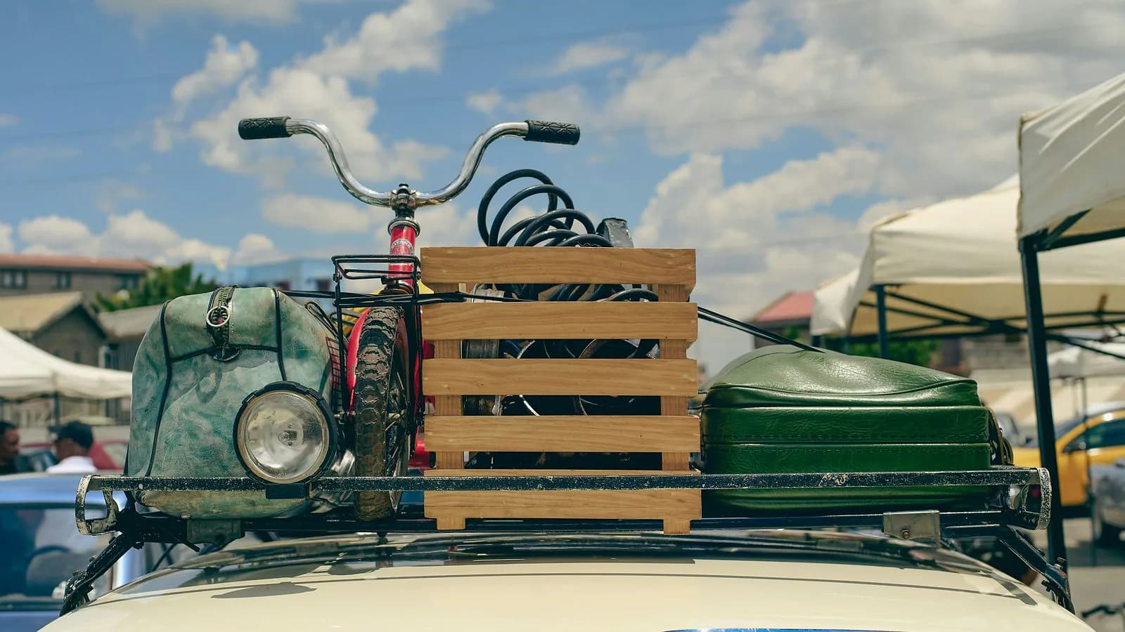 A bicycle, a wooden crate and vintage bags strapped to the roof rack of a cream car under a blue sky with white clouds.