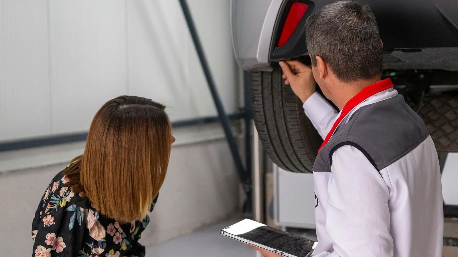 An agent in a work jacket and a customer crouch to inspect the rear wheel of a raised car, the agent holding a tablet.