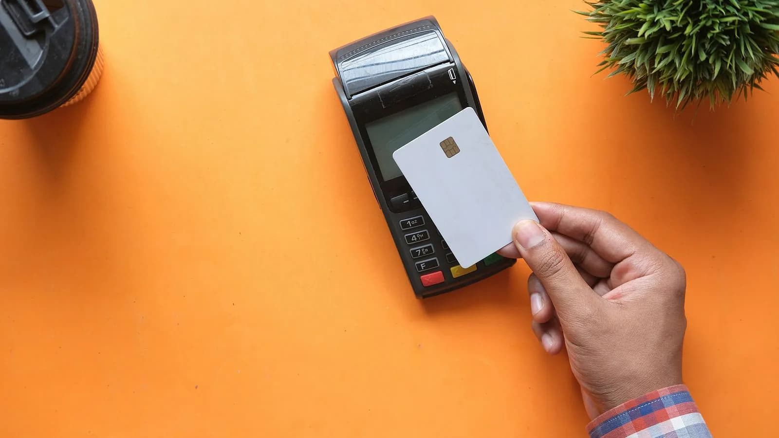 A hand holds a blank white bank card above a payment terminal on a bright orange table, with a small plant in the corner.