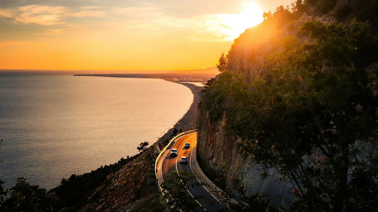 A coastal road winding along a cliff above a calm sea at golden sunset, with two cars driving along the bend.